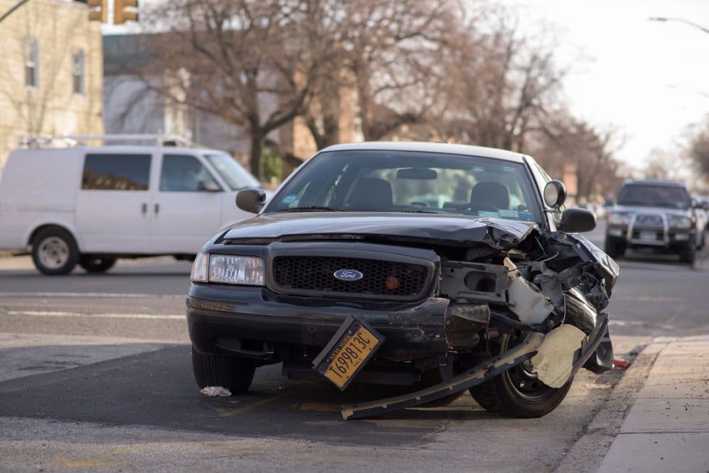 Severely wrecked black Ford sedan with extensive front-end damage and crumpled hood after a serious car accident on a New York street, license plate visible, illustrating high-impact collision injuries