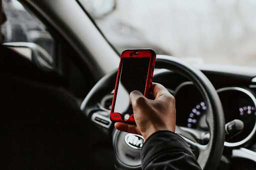 Man holding red smartphone in one hand while gripping steering wheel inside car, illustrating distracted driving and increased risk of car accidents or injuries
