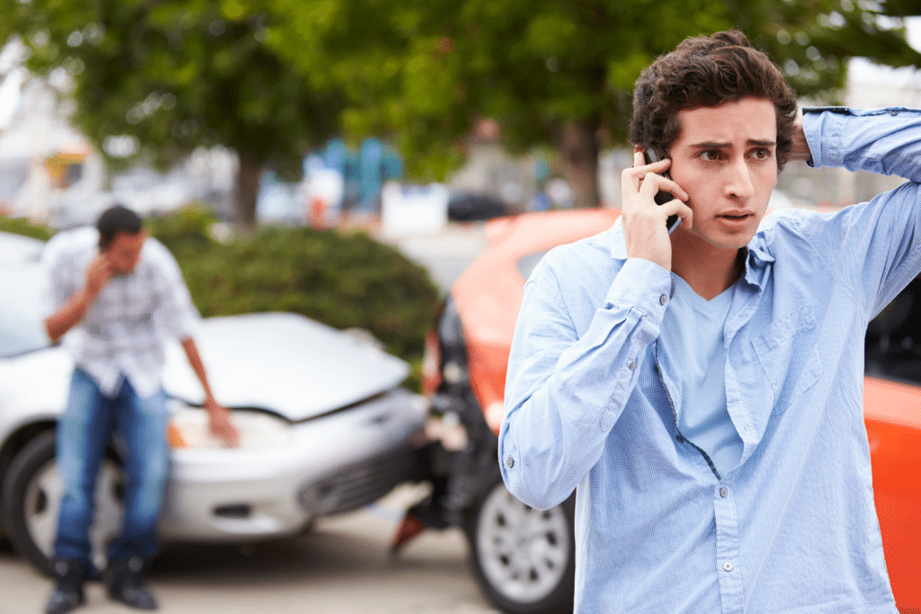 Young man in blue shirt standing stressed outside damaged car after collision, holding phone to ear while another person inspects vehicle damage, typical post-accident scene requiring immediate medical and insurance help