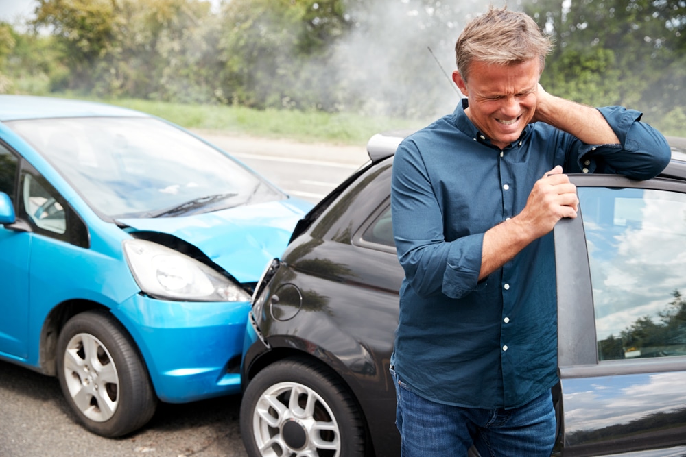 Middle-aged man holding neck in severe pain standing beside damaged blue and black cars after collision on road, showing common whiplash or cervical strain symptoms following car accident