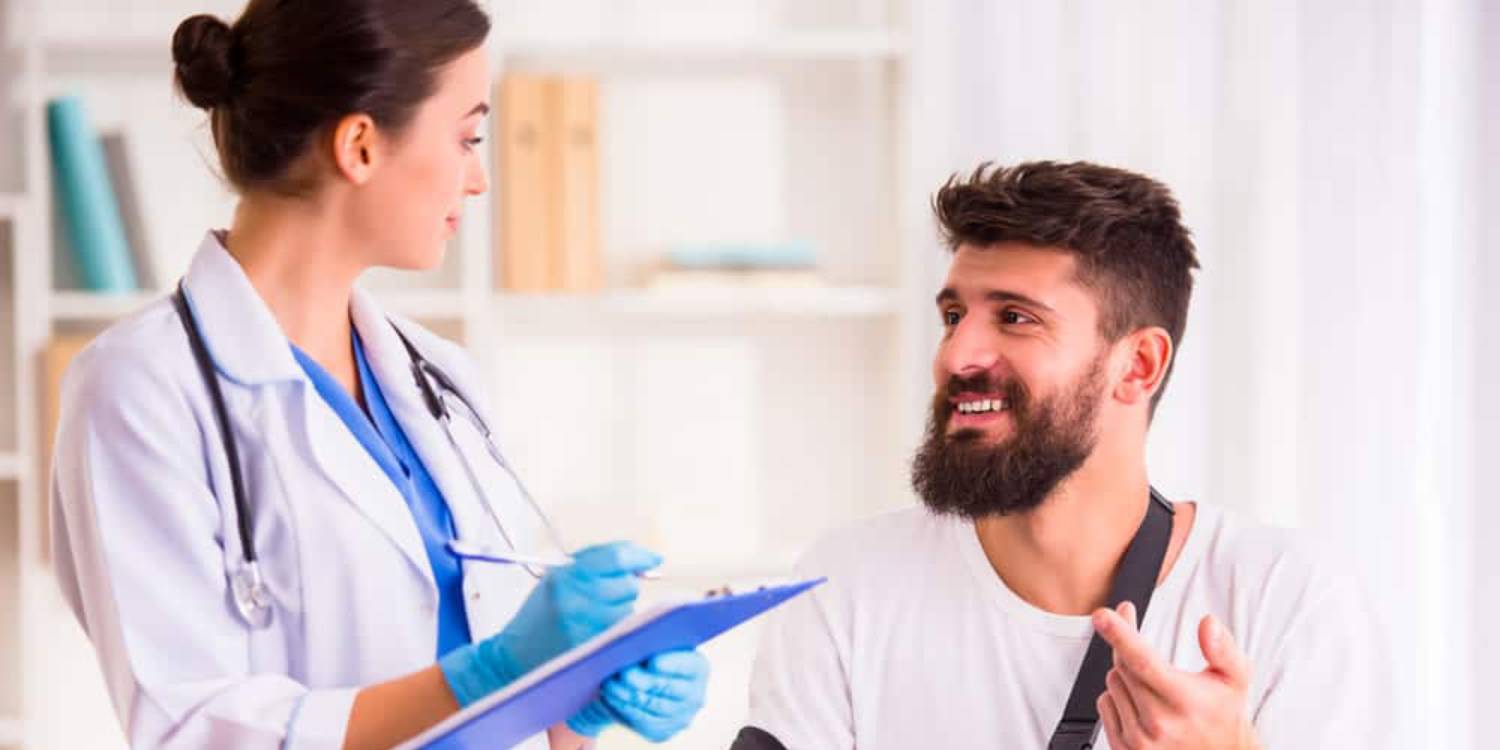 Female doctor in white coat with clipboard discussing treatment plan with smiling bearded male patient after car accident injury in New York clinic