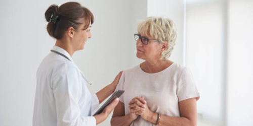 Female doctor in white coat with clipboard gently reassuring senior woman patient during consultation in medical office for injury evaluation or pain management